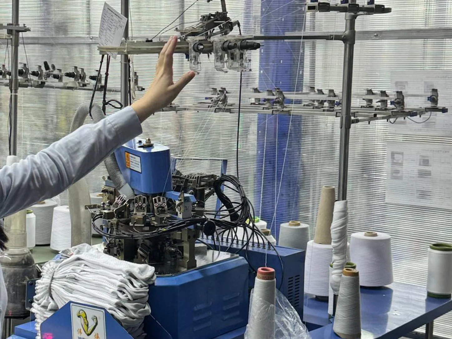 Une machine à tricoter circulaire en cours d'inspection dans une usine chinoise, avec des cônes de fil et des articles tricotés finis à côté